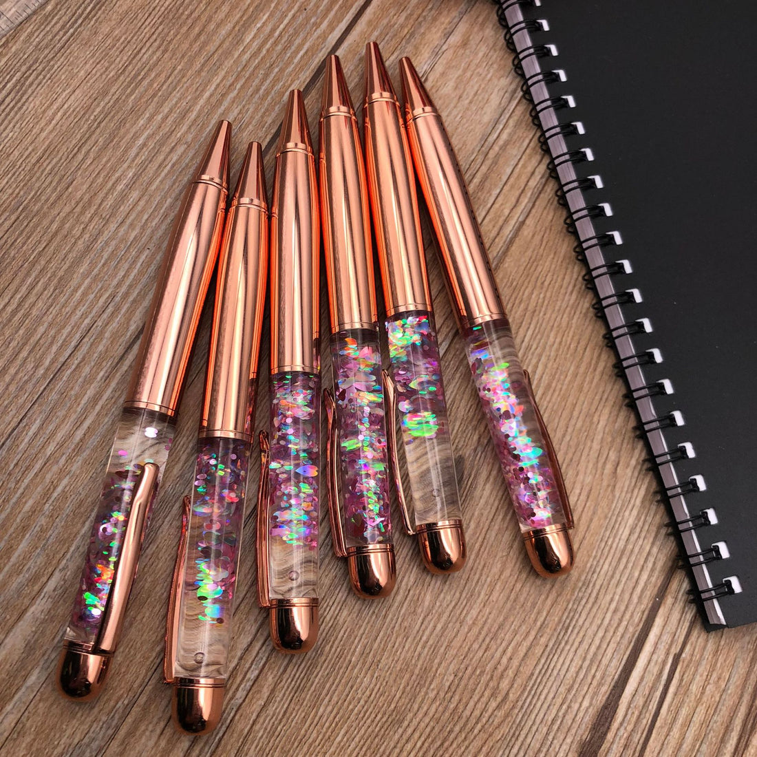Stationery Large Fat Rose Gold Liquid Ball Pen set with shimmering glitter on a wooden surface next to a notebook.