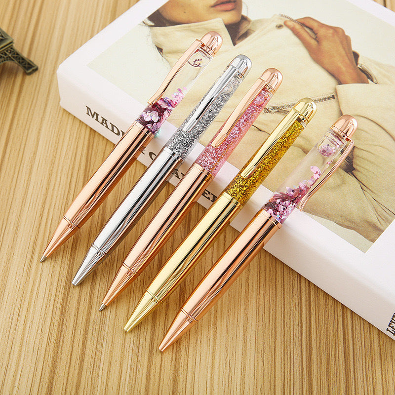 Stationery Large Fat Rose Gold Liquid Ball Pen and three other decorative ballpoint pens on a wooden surface.